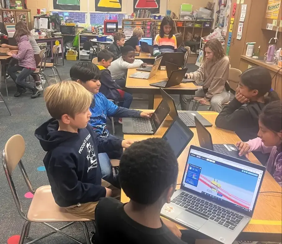 A class of students working on Qweebi projects on their laptops while their teacher sits among them.