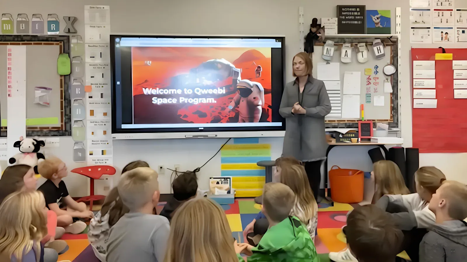 A teacher introducing a Qweebi space project on the classroom display to students seated on the floor.