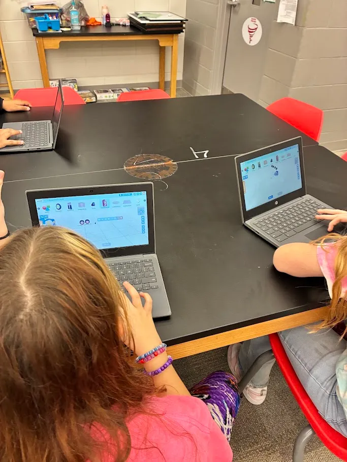 Two students collaborating on a Qweebi project, one helping the other troubleshoot.