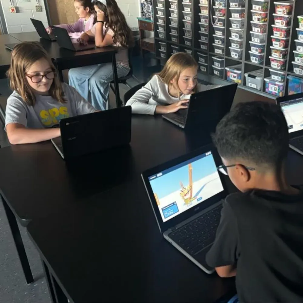 Four students at a shared table building Qweebi projects on their Chromebooks, in front of a wall of classroom supply bins.