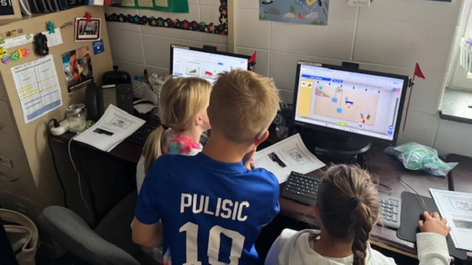 Three students at a bank of desktop computers, working through a Qweebi project with printed worksheets beside them.