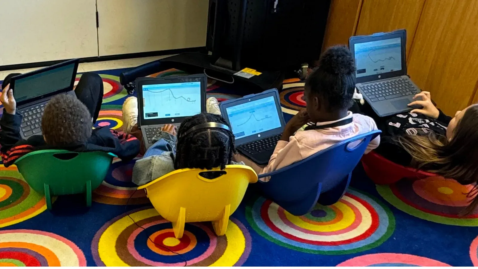 Four young students sitting on floor cushions with Chromebooks on their laps, building Qweebi projects.